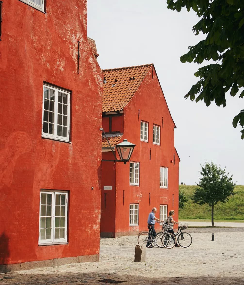 Two people walk with bicycles beside vibrant red buildings on a cobblestone street, under a clear sky with a nearby tree.