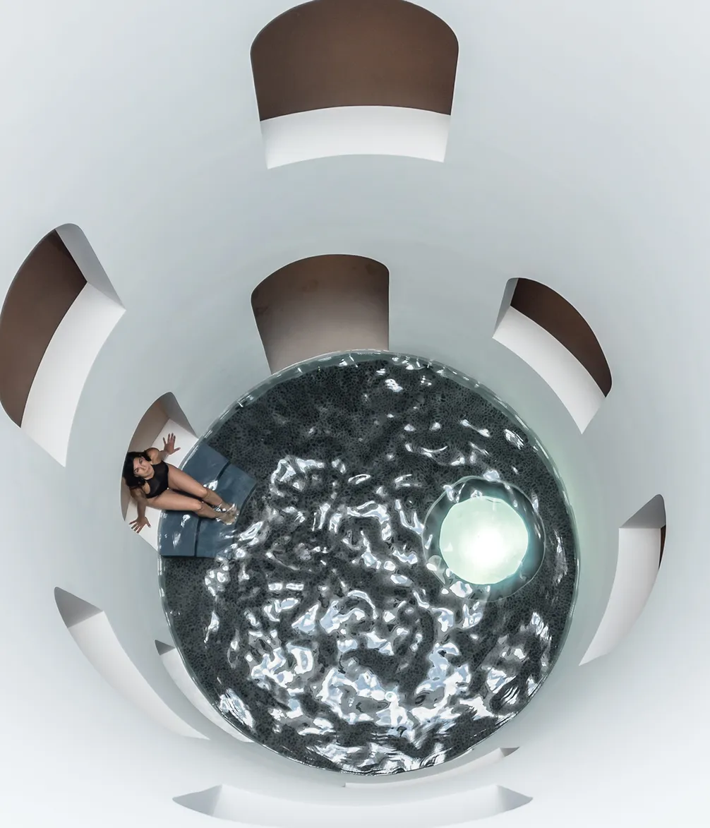 A woman in a black swimsuit sits on a white ledge beside a circular pool with dark water and a glowing orb, surrounded by futuristic white walls.
