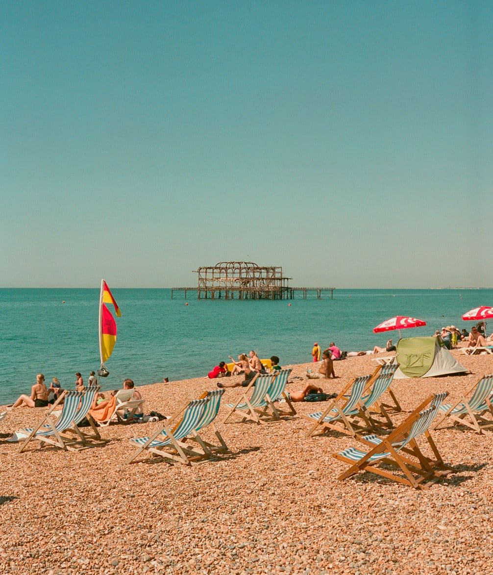 People relaxing on a pebble beach with scattered deck chairs and colorful flags. A rusted pier structure stands in the calm sea under a clear sky.