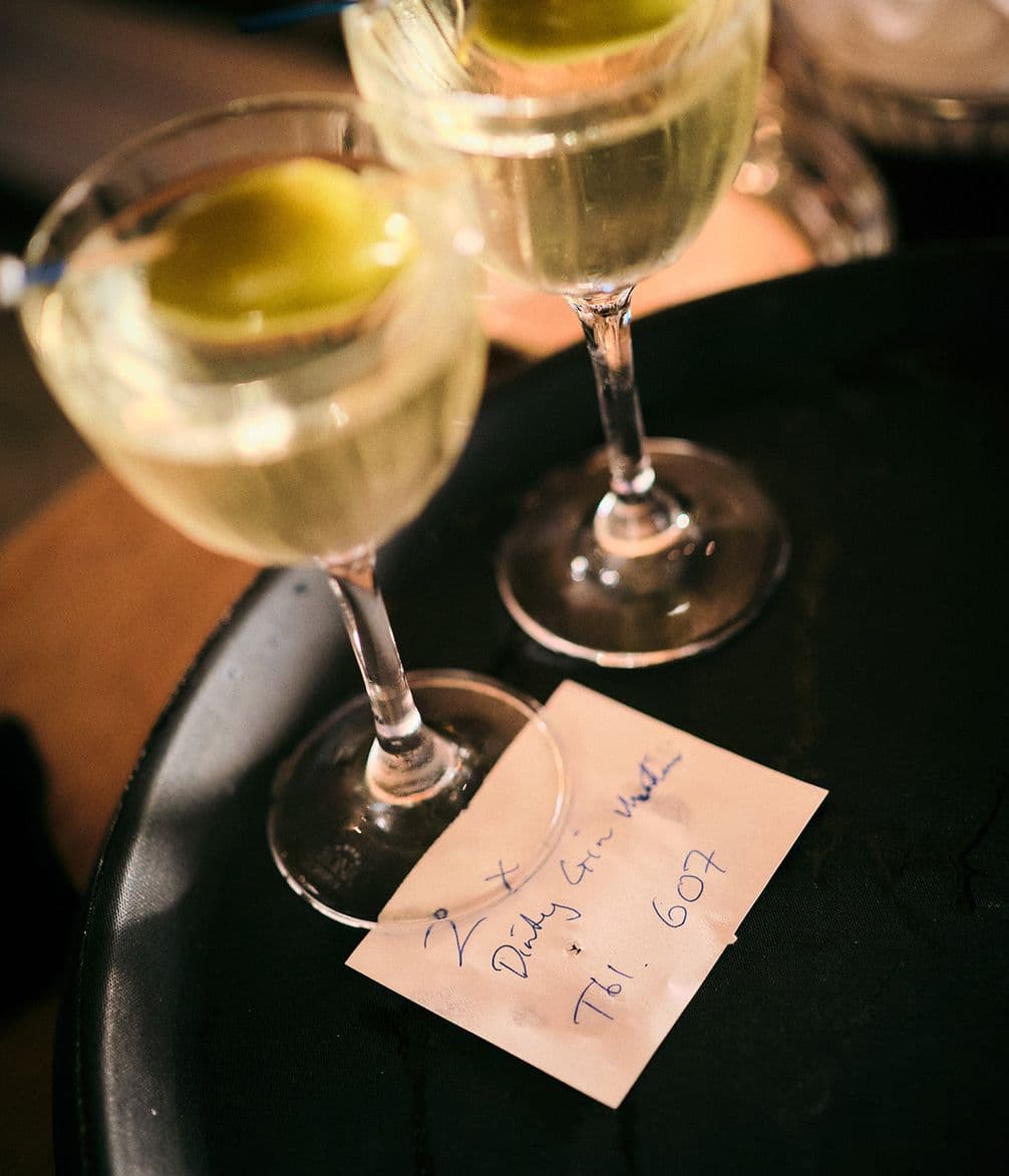 Two cocktails with olives on a tray beside a handwritten note reading "2x Dirty Gin Martinis" and "Tbl: 607."