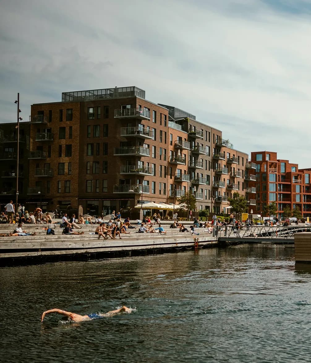 Urban waterfront scene with a swimmer in the foreground and people relaxing on a pier. Brick residential buildings in the background under a cloudy sky.