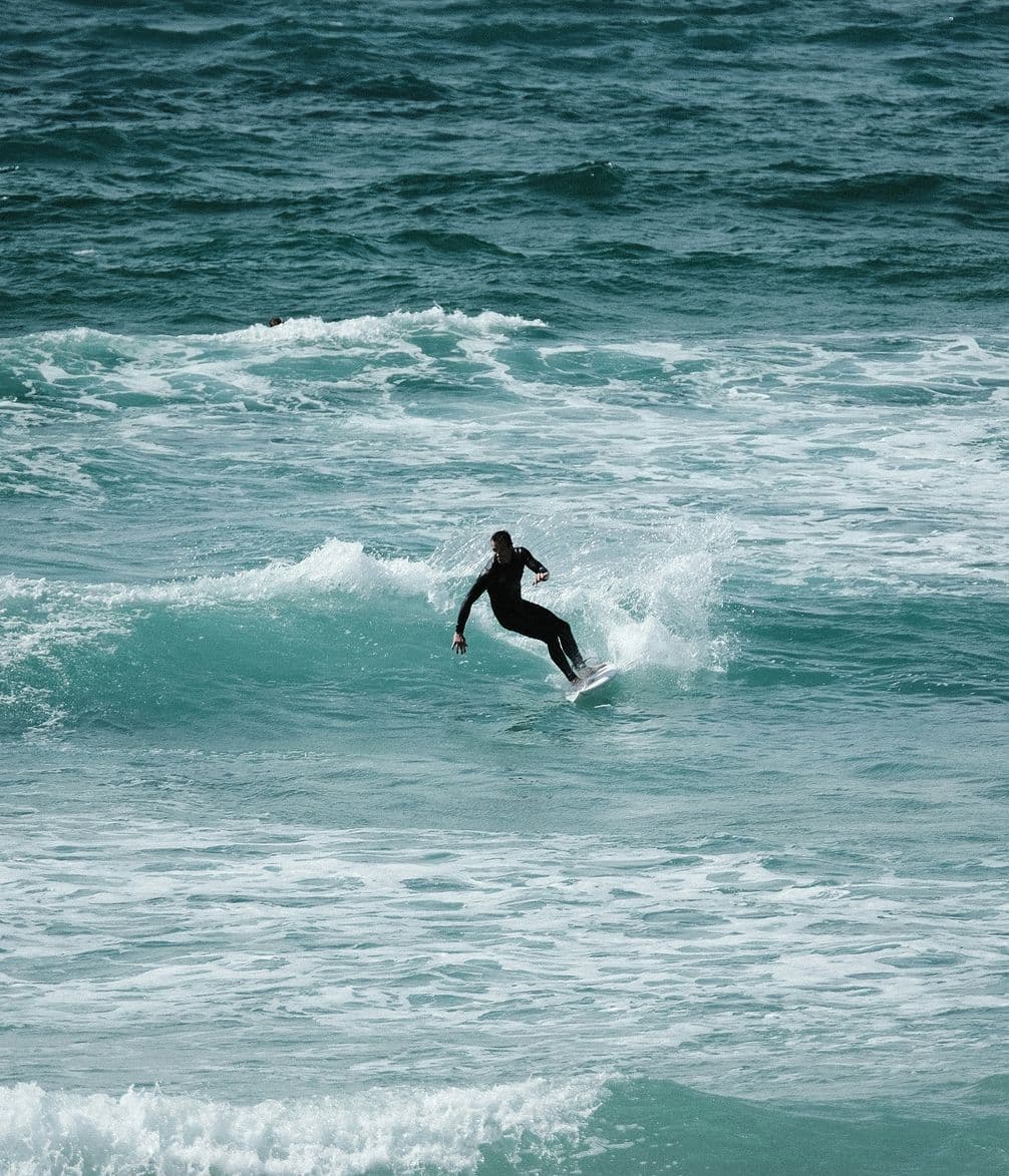 A surfer in a black wetsuit rides a small wave in the ocean under overcast skies, surrounded by choppy, teal-colored water.
