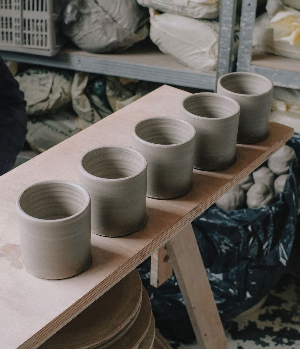 Five unglazed ceramic mugs are lined up on a wooden board in a pottery workshop, with shelves and bags in the background.