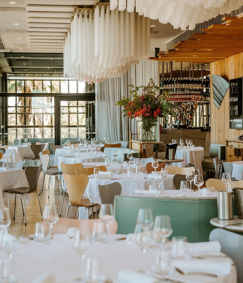 Elegant restaurant interior with white tablecloths, modern chairs, a large floral arrangement, and a bar area in the background.