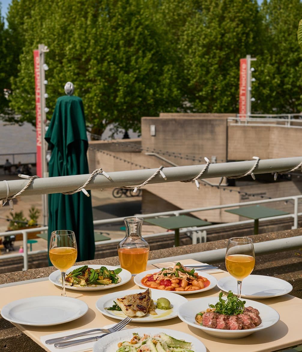 Outdoor dining setup with various dishes and drinks on a table, overlooking a public space with trees and buildings in the background.