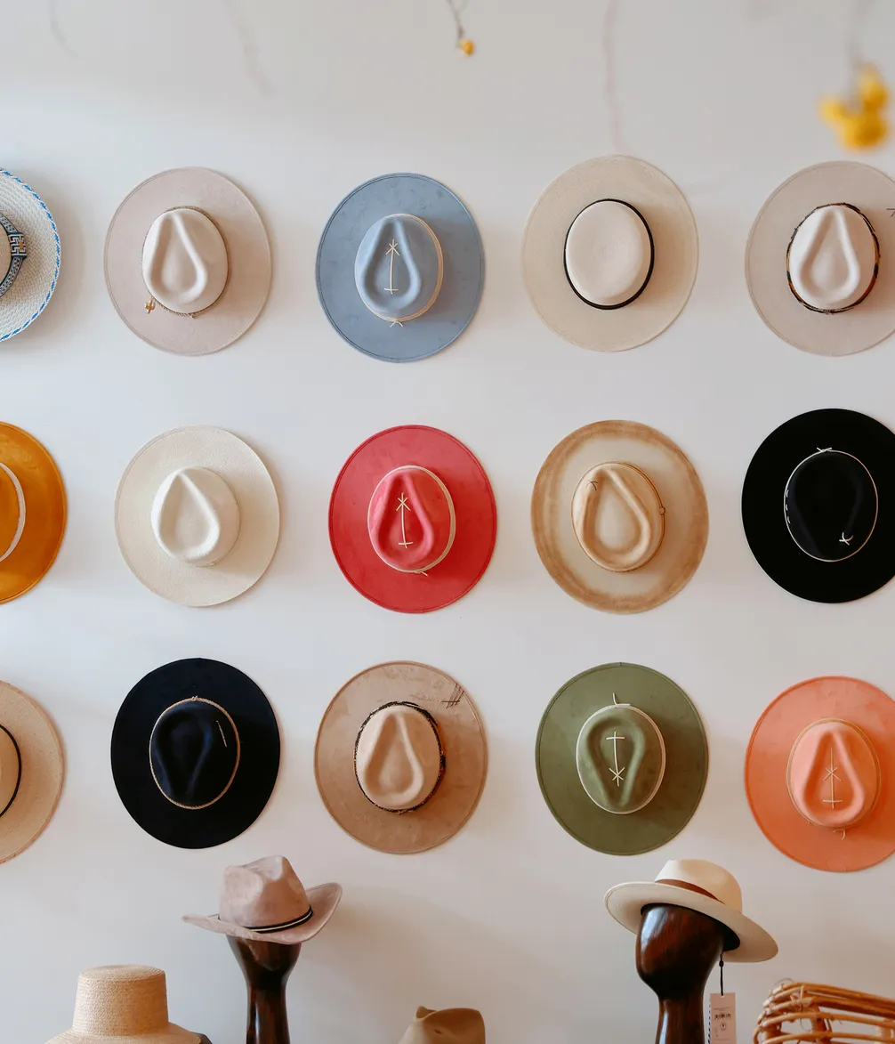 Various colorful wide-brimmed hats hanging in rows on a white wall, with a few displayed below on wooden stands.