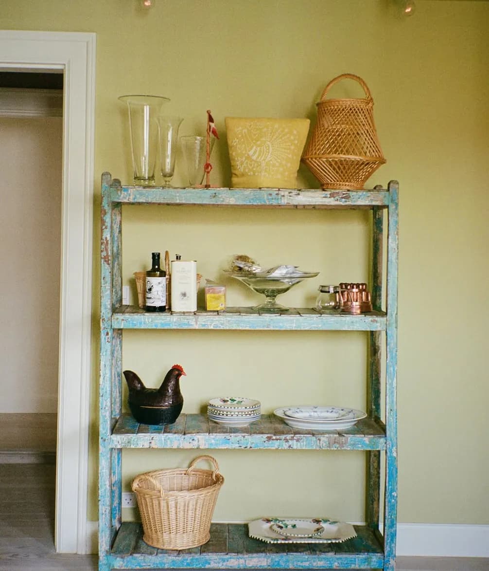 A rustic blue wooden shelf holds glass vases, a woven basket, a decorative bowl, a ceramic rooster, tins, and kitchen items against a pale green wall.