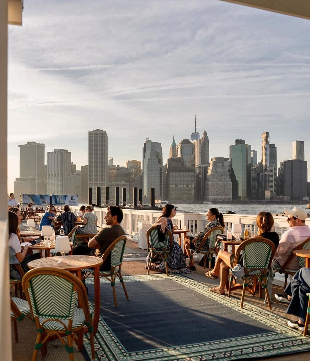 People dining at an outdoor terrace with a view of a city skyline across the water, under a partly cloudy sky.