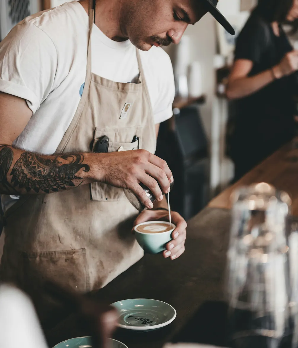 Barista with a tattooed arm in a beige apron prepares a cup of coffee, using a spoon to stir in a cozy cafe setting.