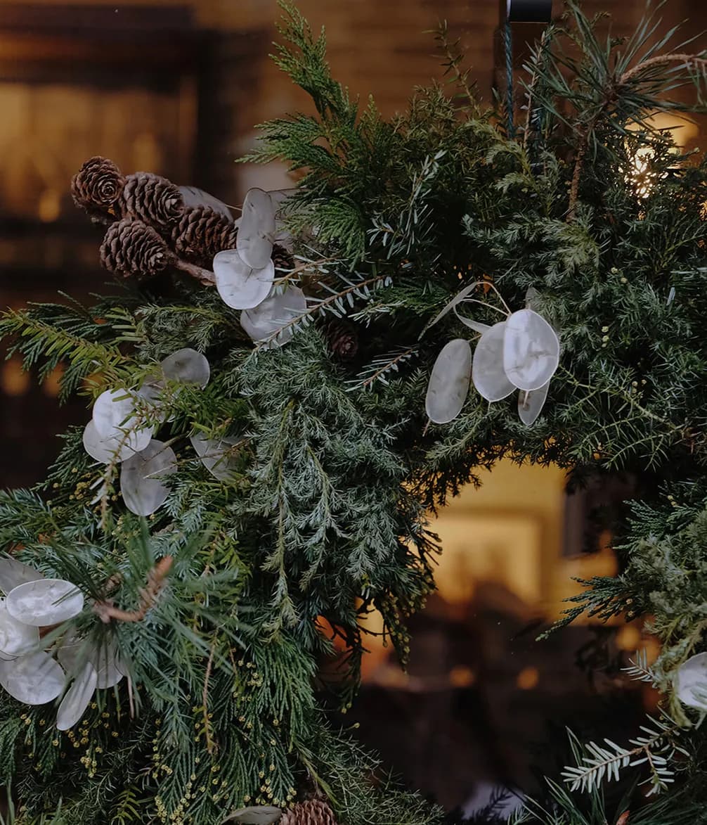 Festive wreath with pinecones, green foliage, and translucent white leaves, hanging against a blurred indoor background.