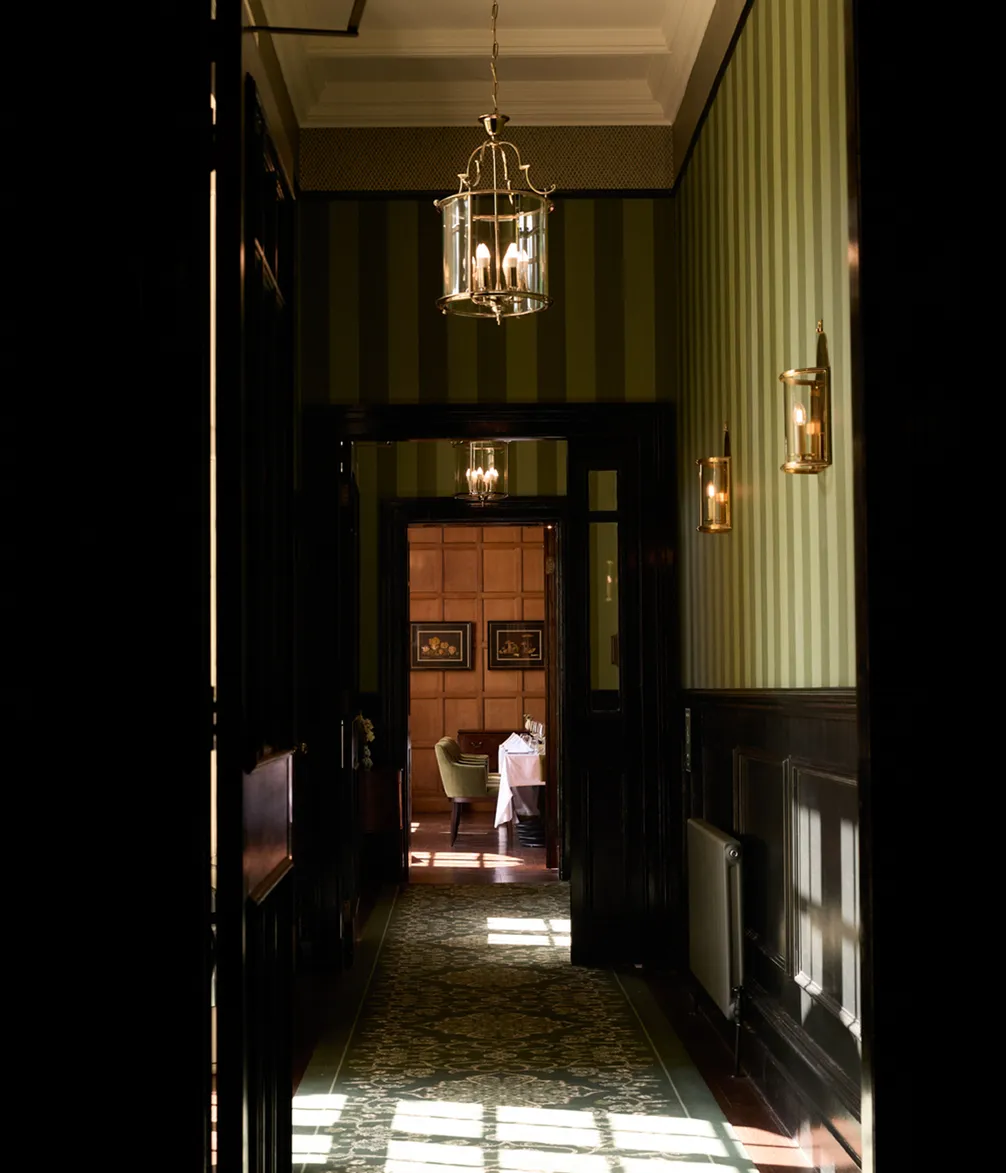 A dimly lit hallway with striped walls, leading to a cozy room with a table set for dining. Centered hanging and wall lights cast a warm glow.