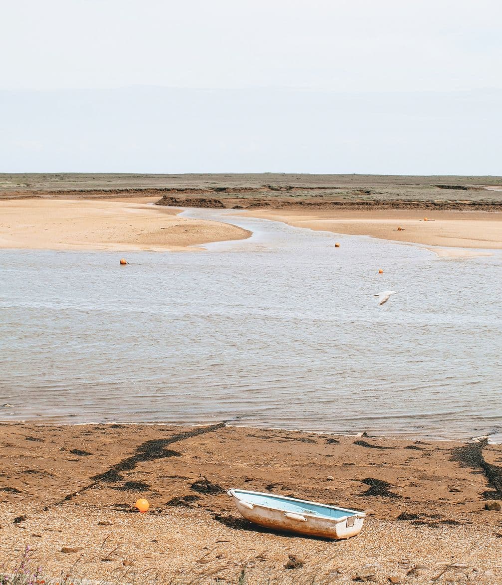 A small boat rests on a sandy beach beside a calm river, with orange buoys dotting the water and a vast, flat landscape in the background.