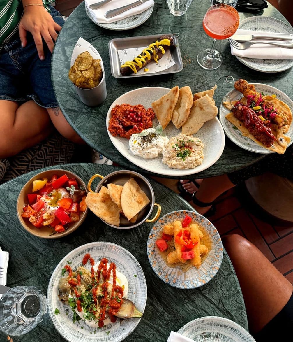 A table with various Mediterranean dishes including dips with pita, grilled meat, salad, and a hot dog, alongside a pink cocktail.