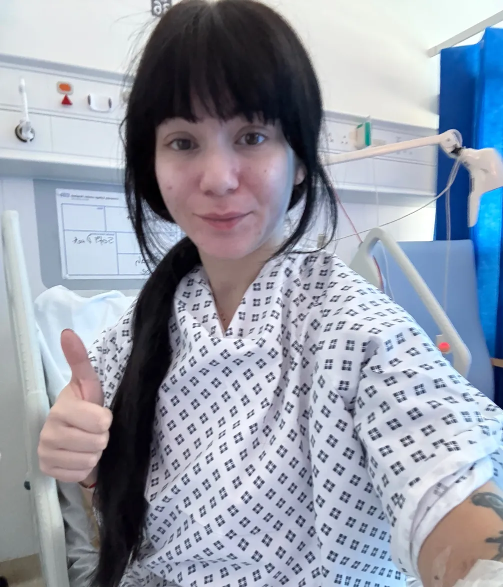 A young woman with dark hair, wearing a hospital gown, smiles and gives a thumbs-up from a hospital bed, appearing to be recovering well.