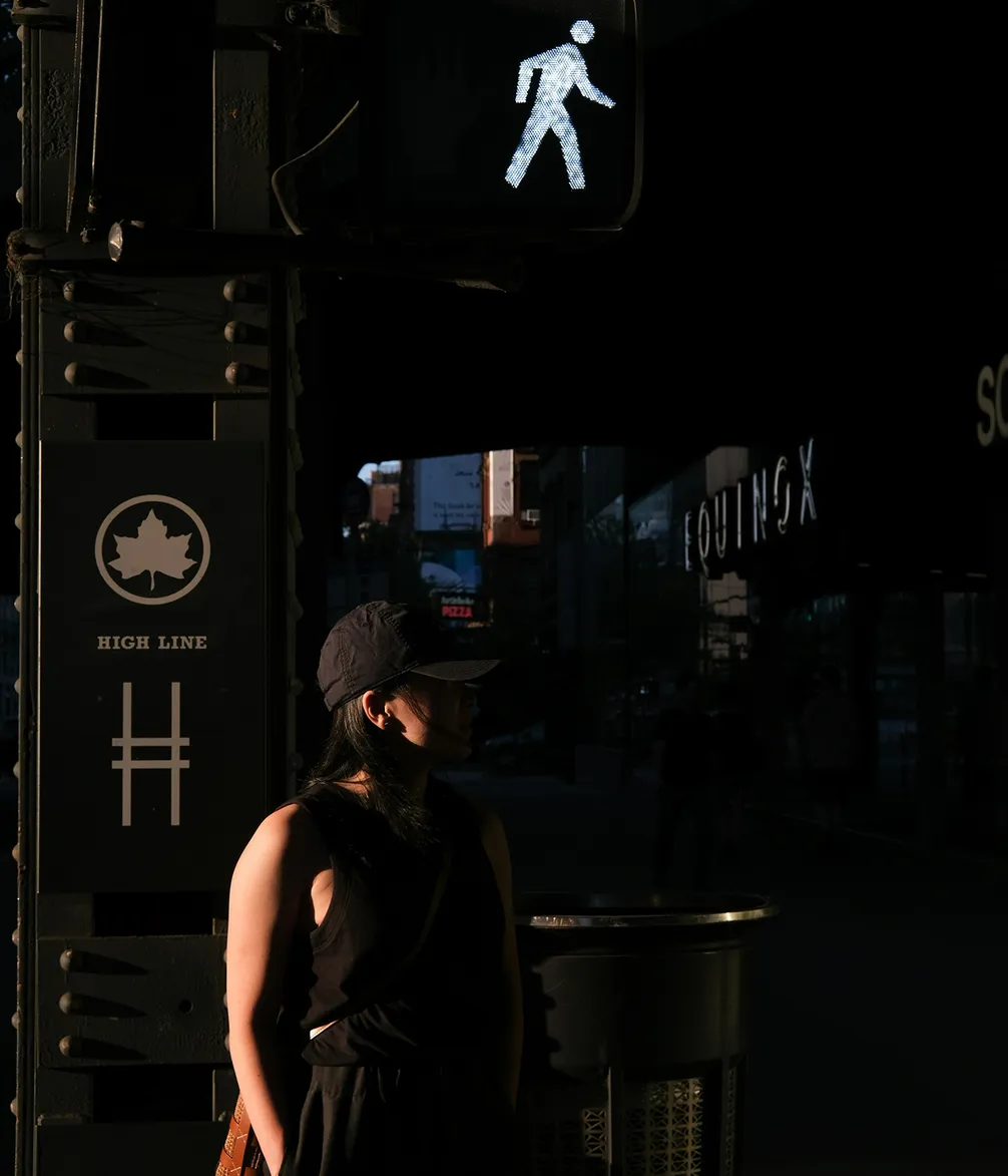 A person in a hat stands in shadow near a pedestrian signal on a city street, with the "High Line" sign and a nearby Equinox gym visible.
