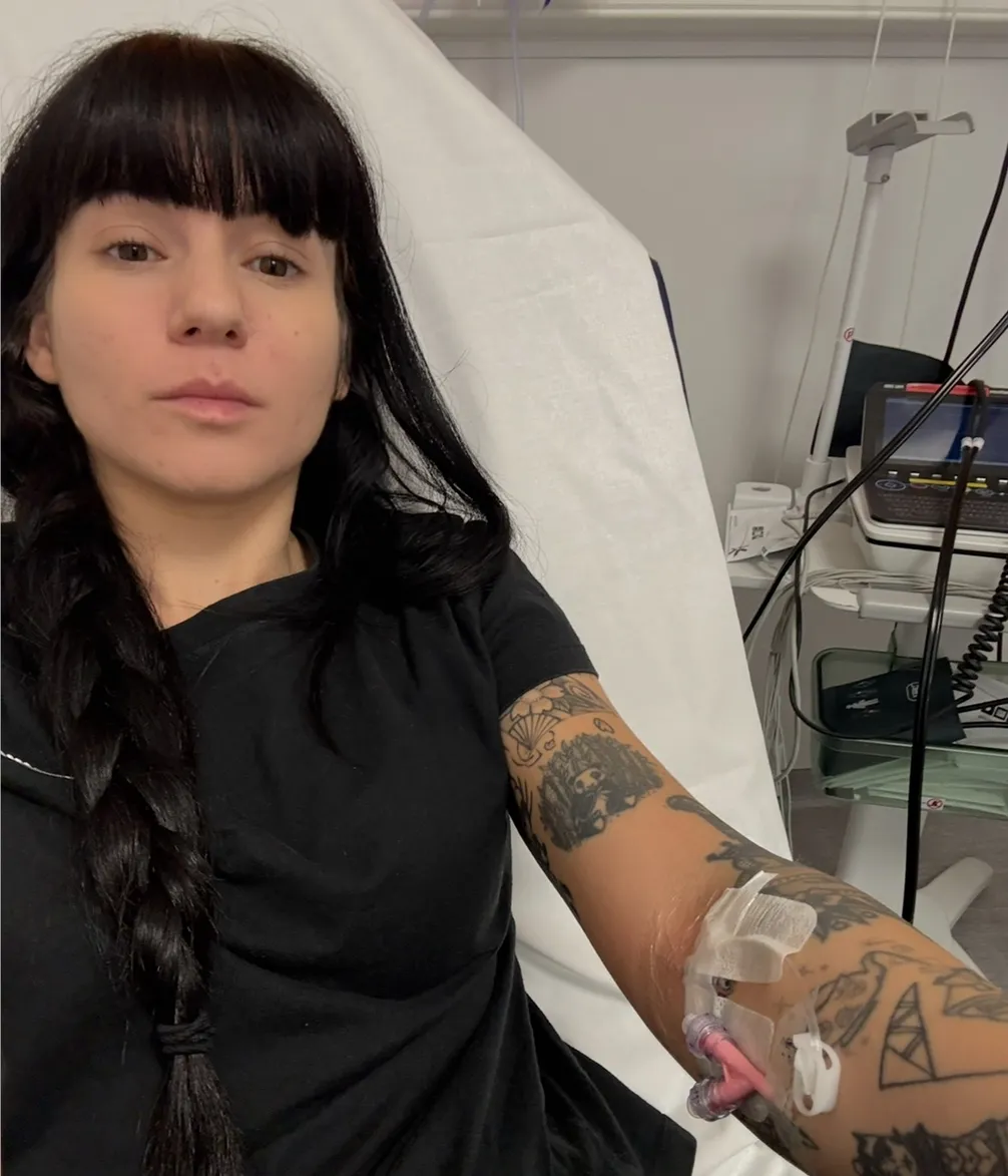 A woman with dark hair and bangs sits in a hospital bed with an IV in her tattooed arm, looking calmly at the camera.