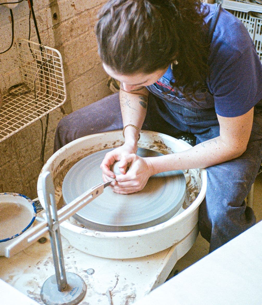 A person with long hair works on shaping clay on a pottery wheel, focusing intently on the clay, in a workshop setting.