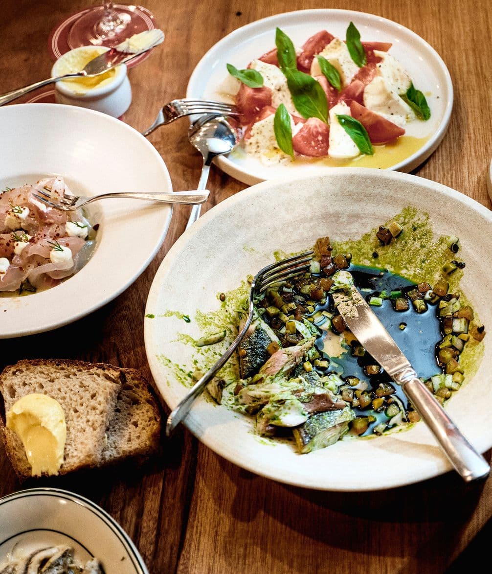 A rustic table setting with plates of salad, fish with sauce, bread with butter, and a small dessert, showcasing various fresh and colorful dishes.