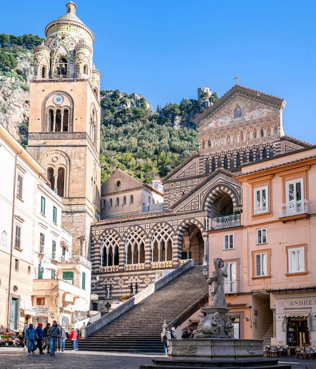 Historic cathedral with a tall bell tower and intricate facade, surrounded by colorful buildings and a stone staircase under a clear blue sky.