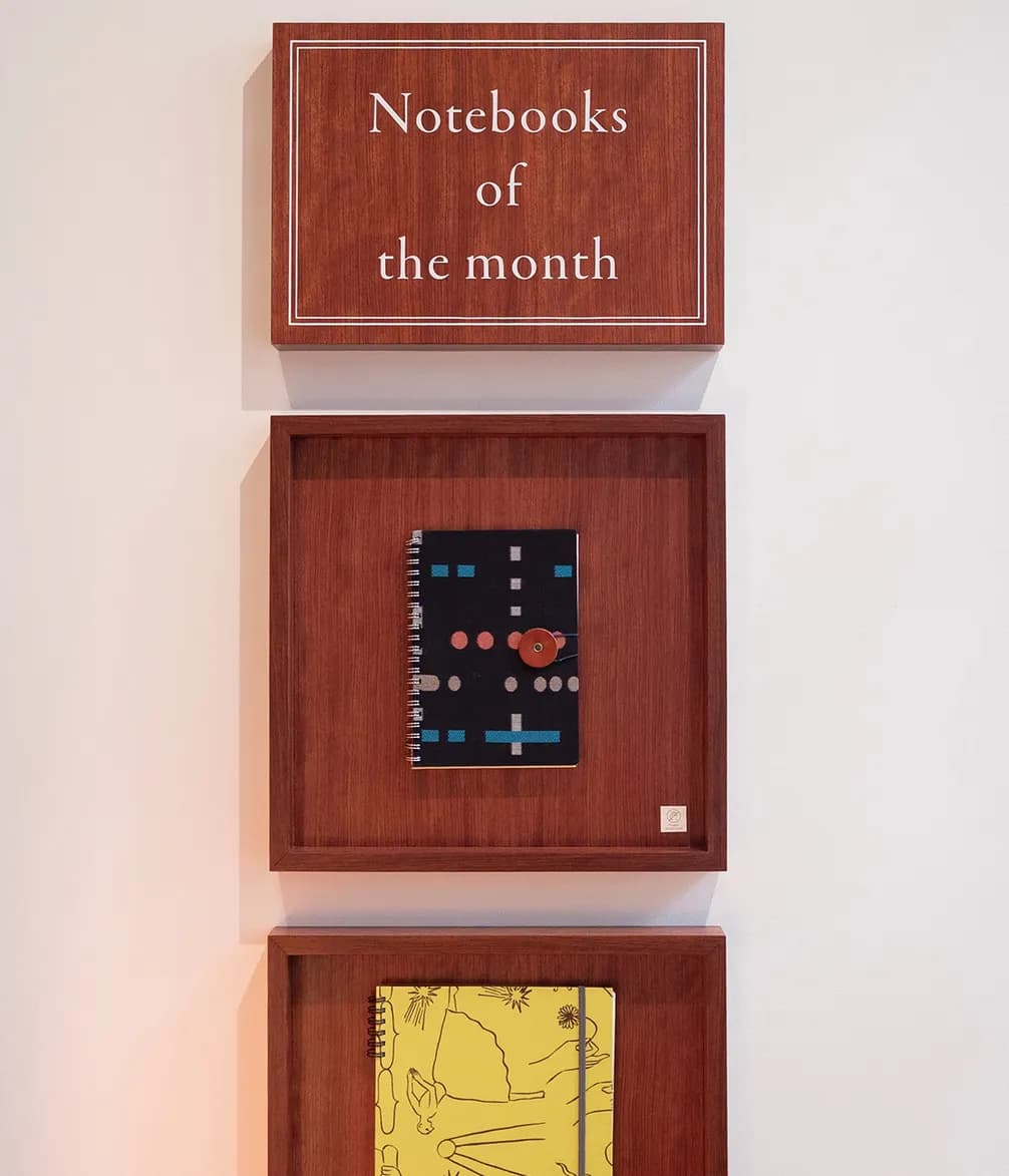 A sign "Notebooks of the month" above two framed notebooks on a white wall; one black with abstract patterns, one yellow with line art.