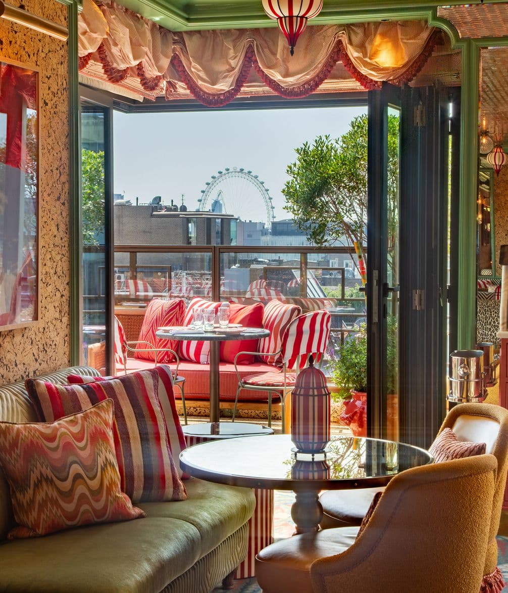 Chic indoor seating area with colorful cushions, open doors to a balcony with red-striped furniture, and a view of an observation wheel in the distance.