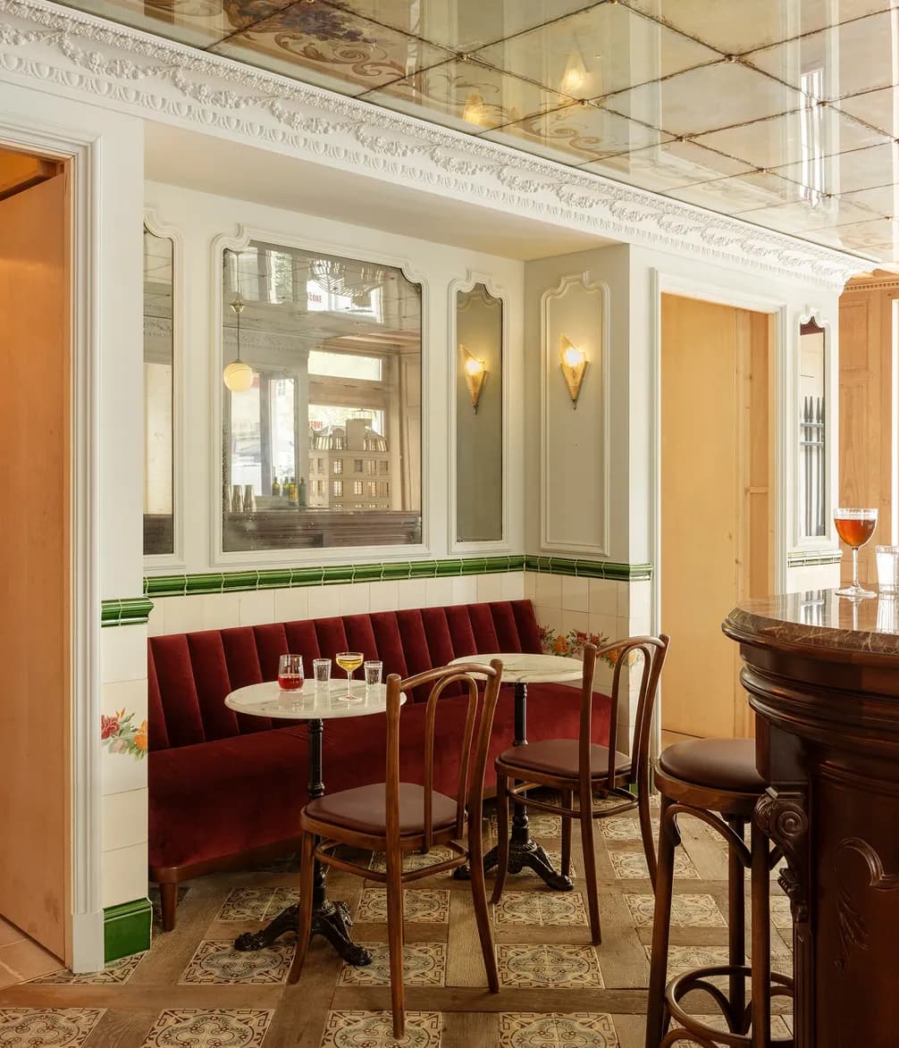 A stylish café interior with vintage decor, featuring a red velvet booth, wooden chairs, marble tables, and a mirrored wall under a decorative ceiling.