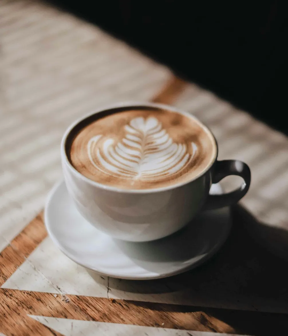 A cup of cappuccino with intricate latte art in the shape of a leaf on top, placed on a wooden table.