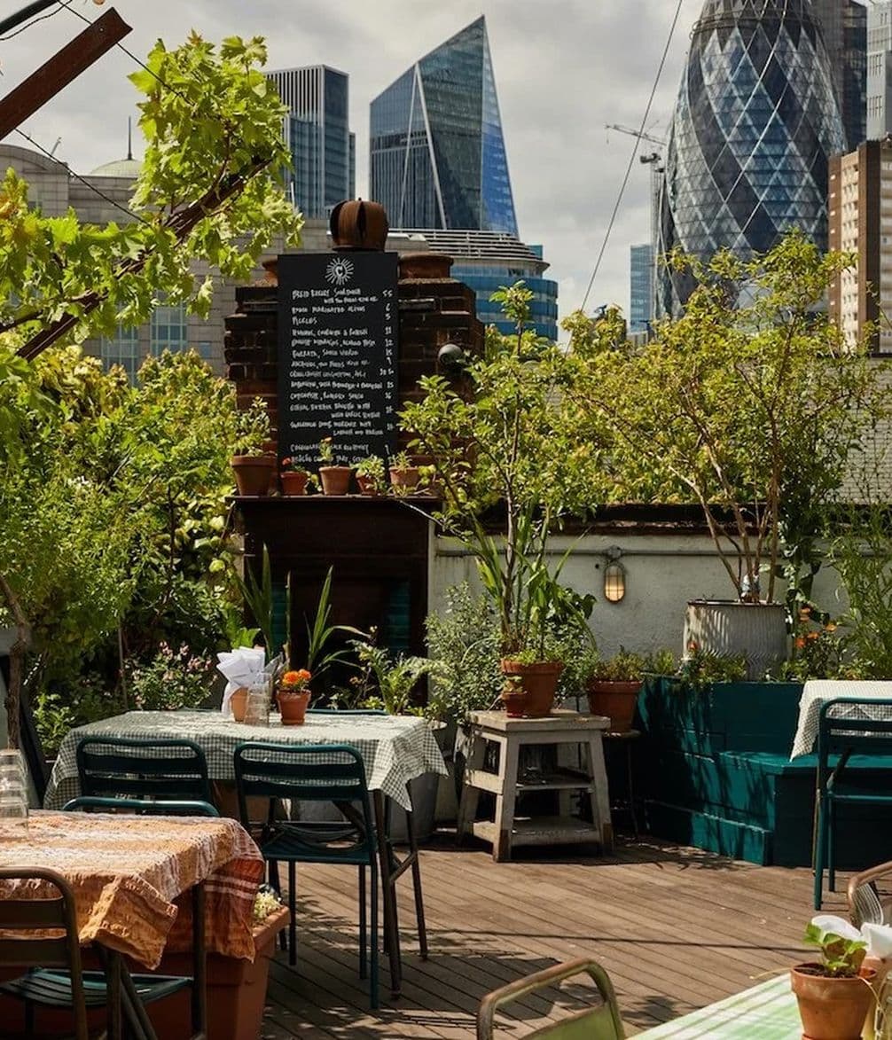 Rooftop garden café with tables, chairs, plants, and skyscrapers in the background under a cloudy sky.