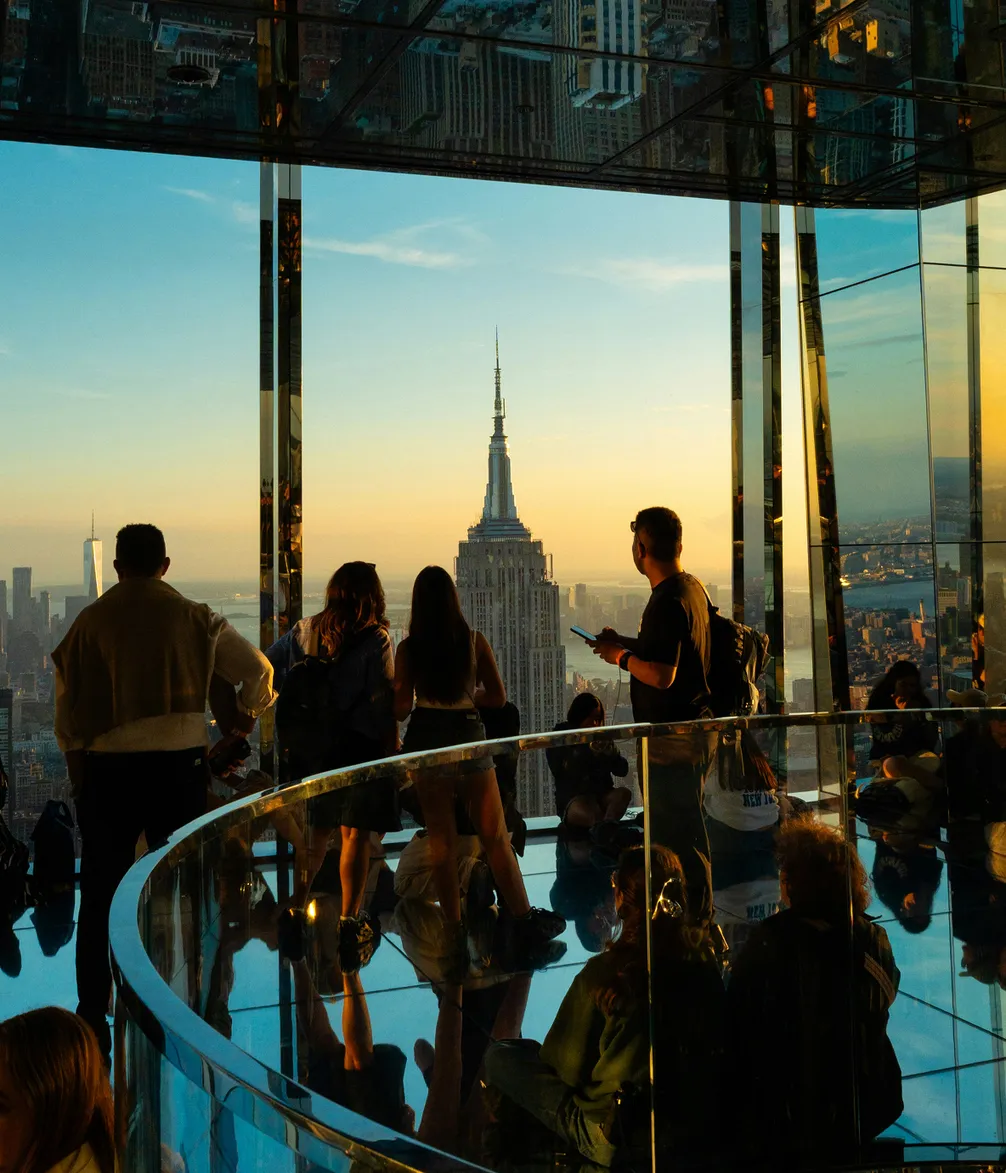 People stand on a glass observation deck with reflections, viewing the Empire State Building at sunset.