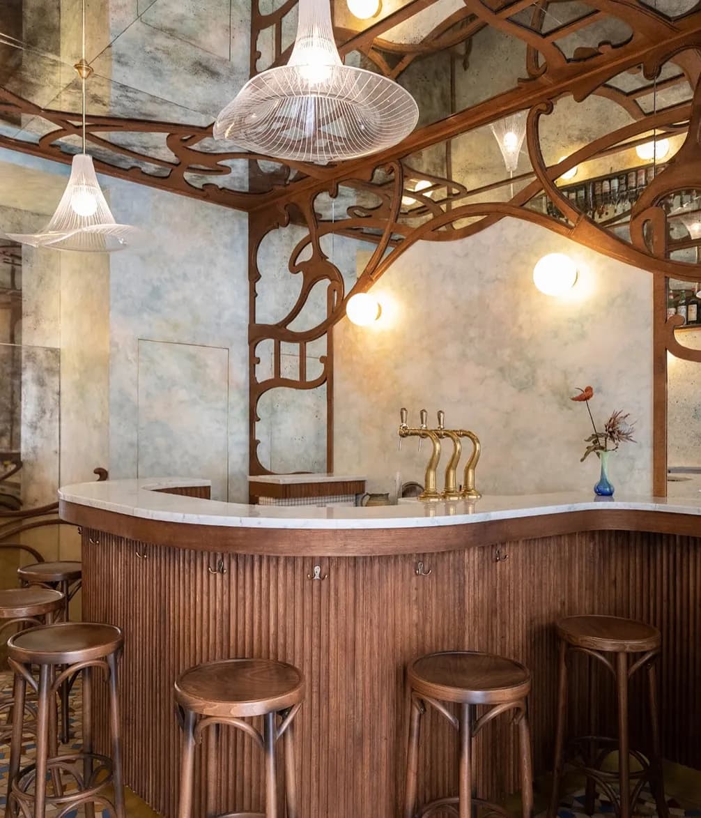 A stylish bar interior with wooden stools, decorative wooden panels, brass taps, and pendant lights, featuring a vase with flowers on the counter.