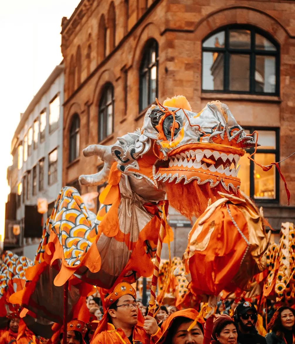 Dragon dance performers in vibrant orange costumes parade through a city street, surrounded by a cheering crowd.