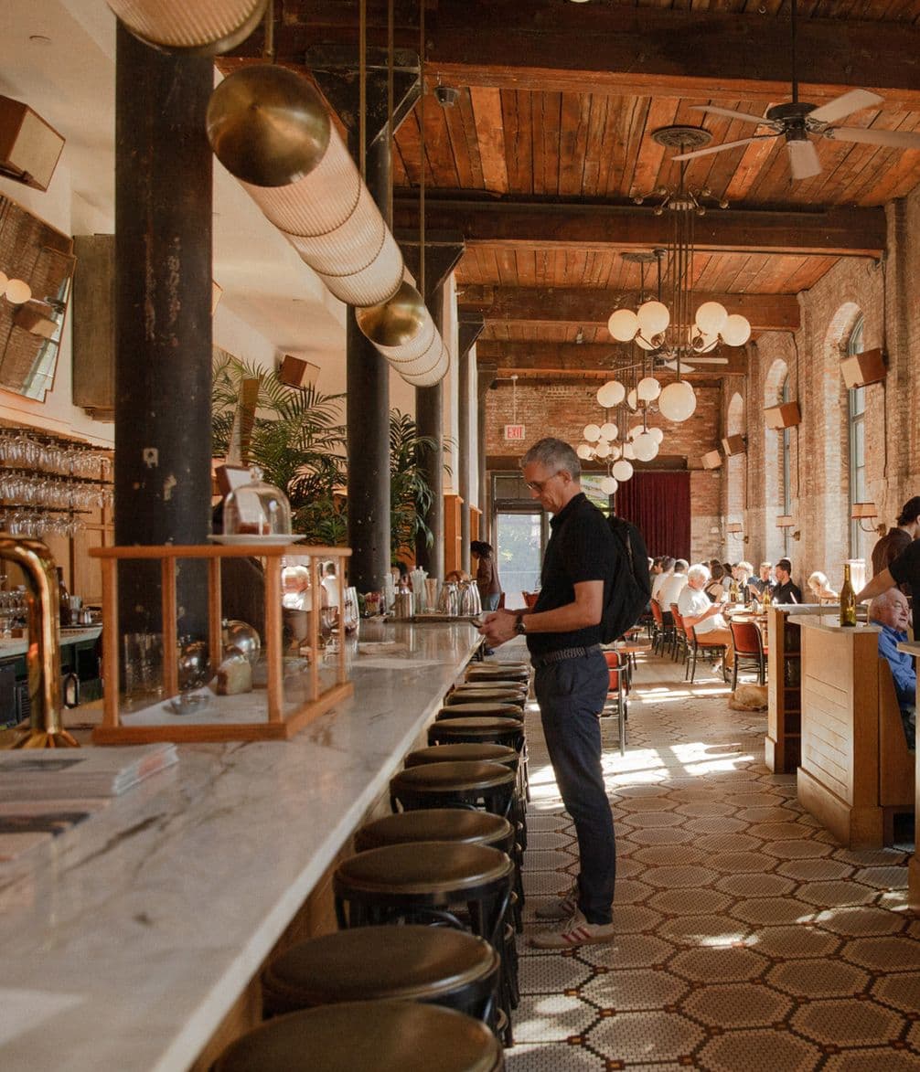 A man stands at a long marble bar in a warmly lit, rustic restaurant with exposed brick walls and large windows.