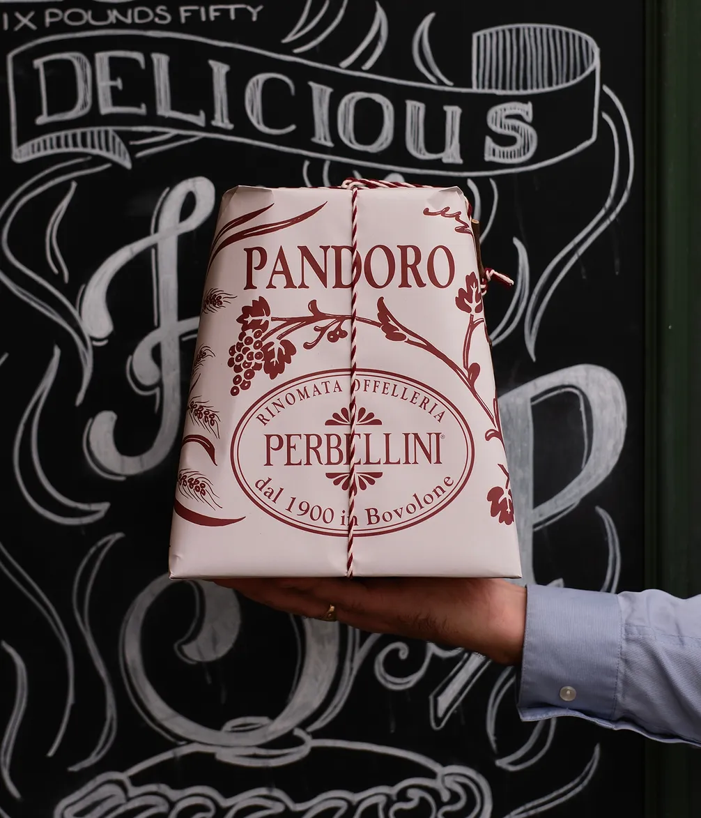 A person holds a wrapped Pandoro cake from Perbellini in front of a chalkboard. The package is decorated with red designs and twine.