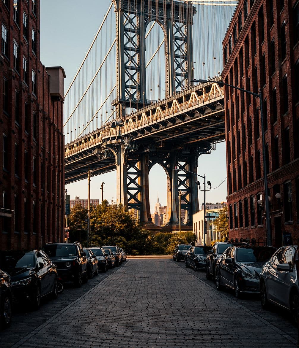 Street view of the Manhattan Bridge framed by red brick buildings, with cars parked along the side and the Empire State Building in the distance.