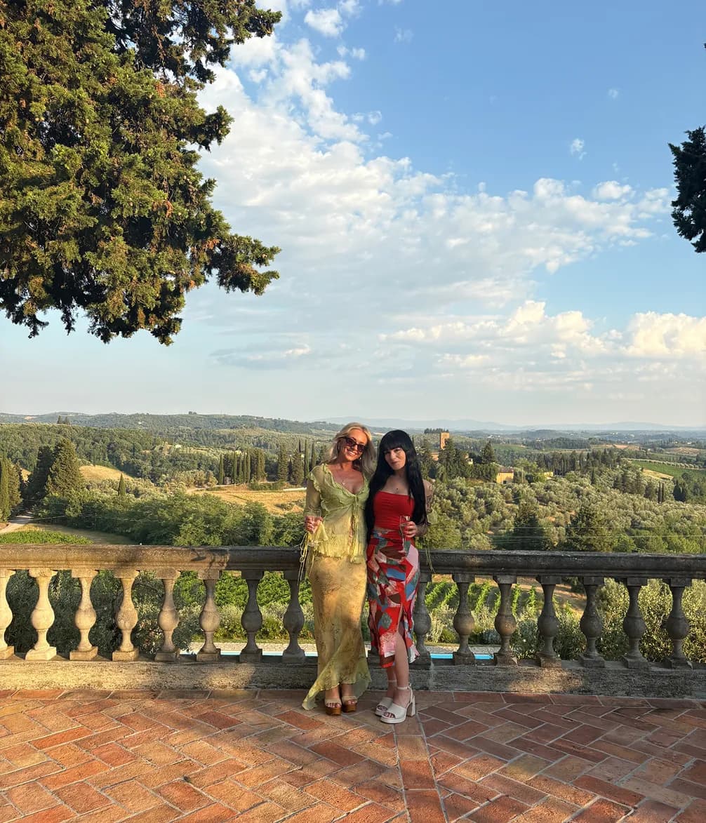Two women on a balcony overlook a sunny Tuscan landscape of rolling hills, vineyards, and olive groves. They hold drinks.