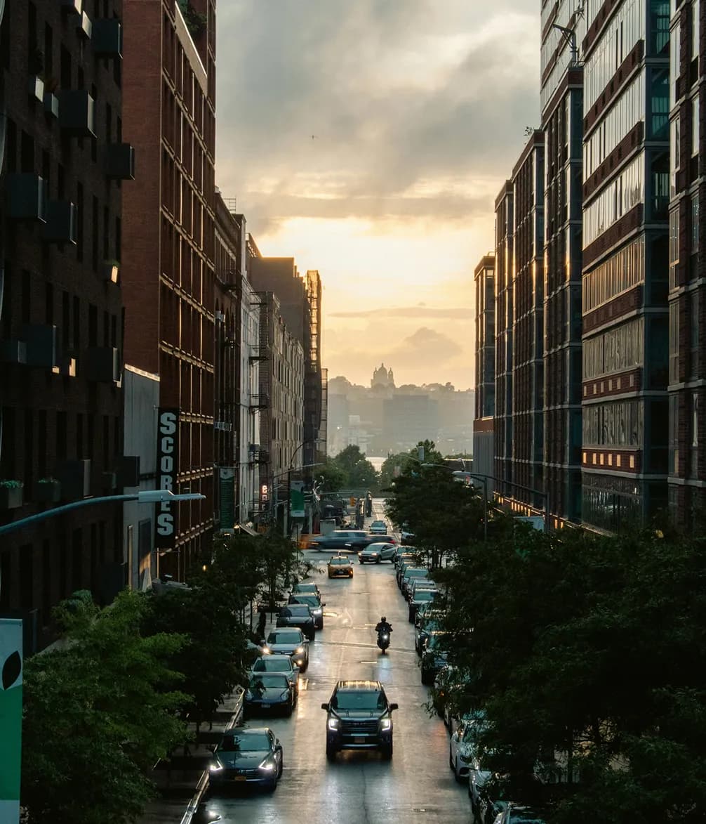 City street at sunset with cars and a motorbike, flanked by tall buildings, and a distant view of a dome against a cloudy sky.