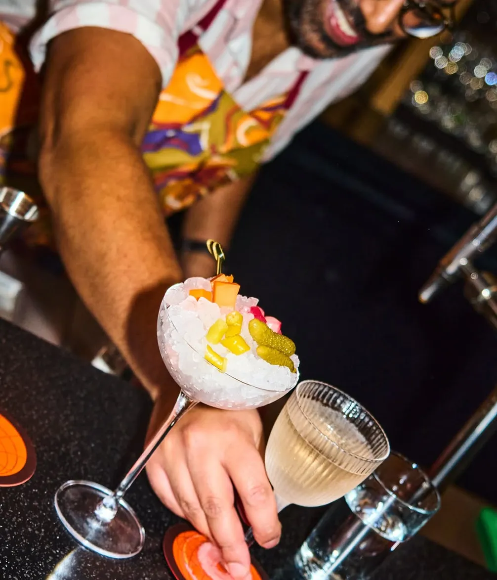 A person in a colorful apron serves a cocktail glass filled with ice, garnished with pickles and candies, beside a small glass of clear liquid.