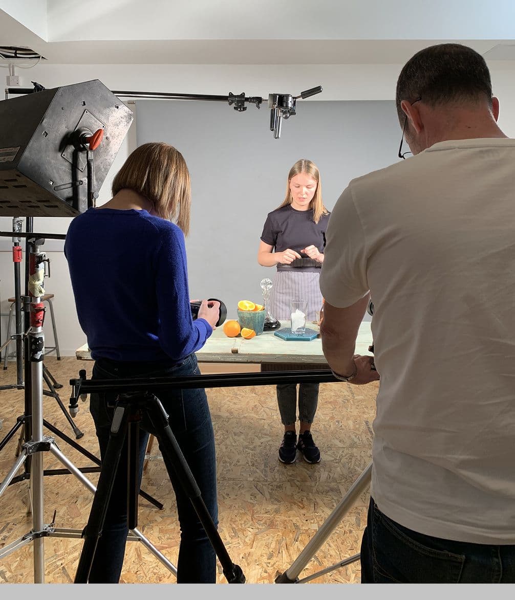 A behind-the-scenes photo shoot with a woman preparing food on a table, surrounded by cameras and lighting equipment, with two crew members.