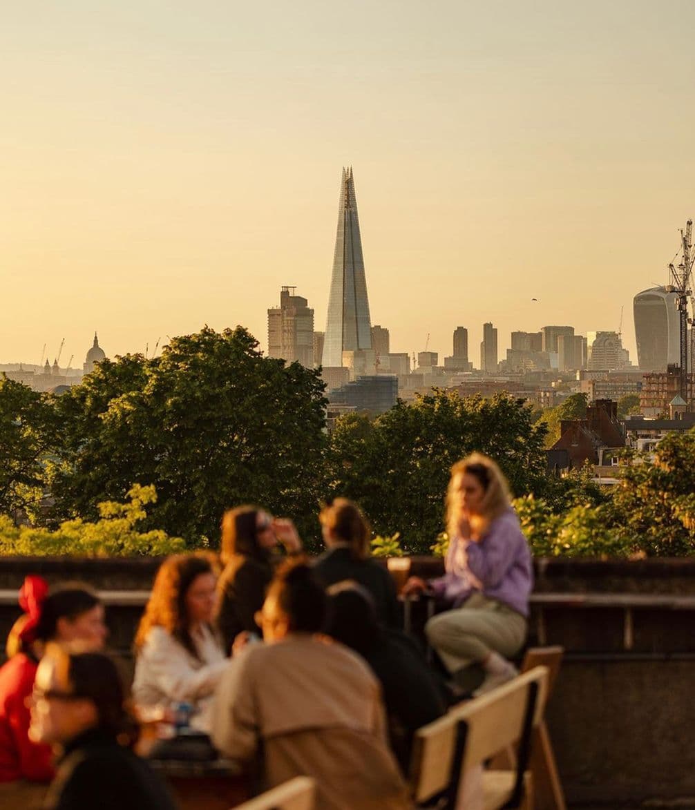 People sitting at an outdoor cafe with a view of London's skyline, including The Shard, during sunset. Lush green trees in the foreground.