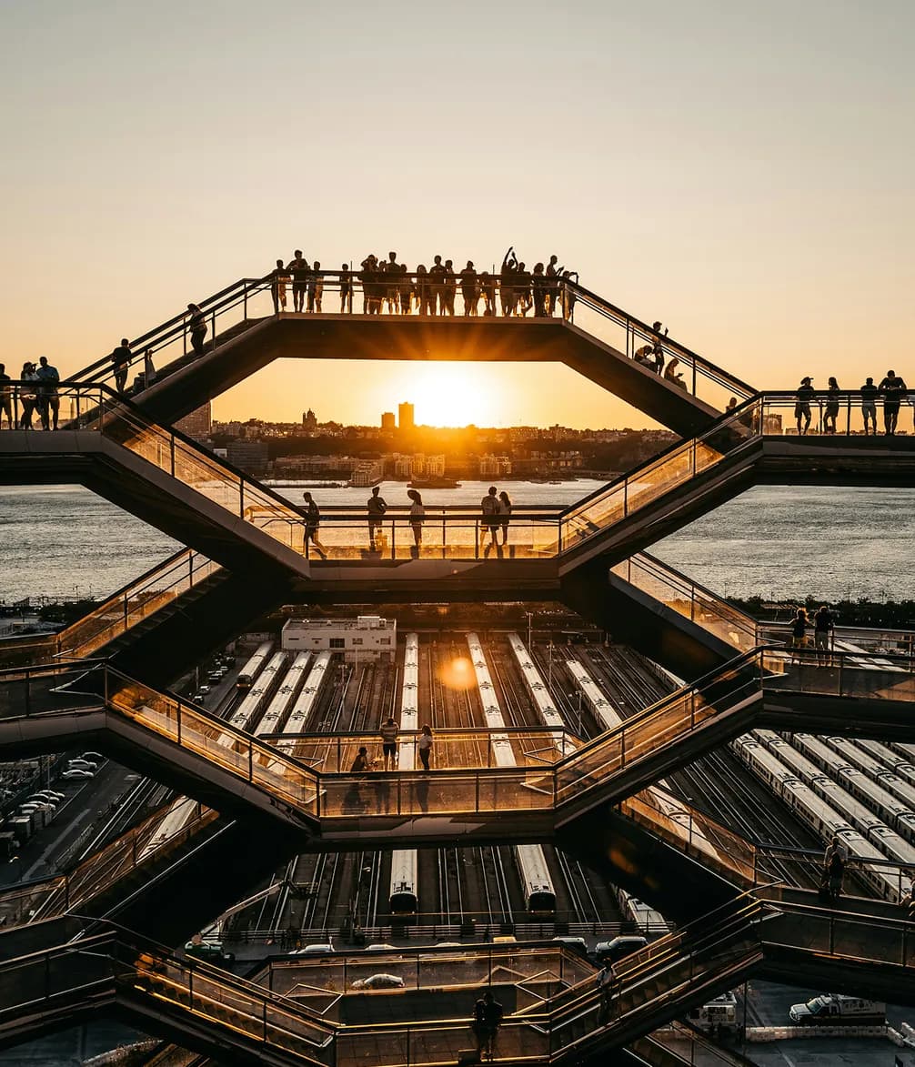 People on a multi-level structure at sunset overlooking train tracks and a river, with the sun setting behind a distant city skyline.