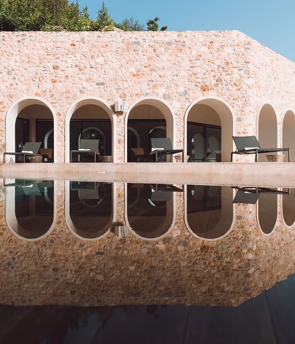 A stone building with arched openings and lounge chairs reflects in a tranquil infinity pool under a clear blue sky.