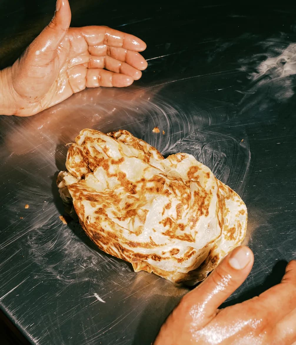 Hands with oil resting on a metal surface beside a freshly made, golden brown roti or flatbread.
