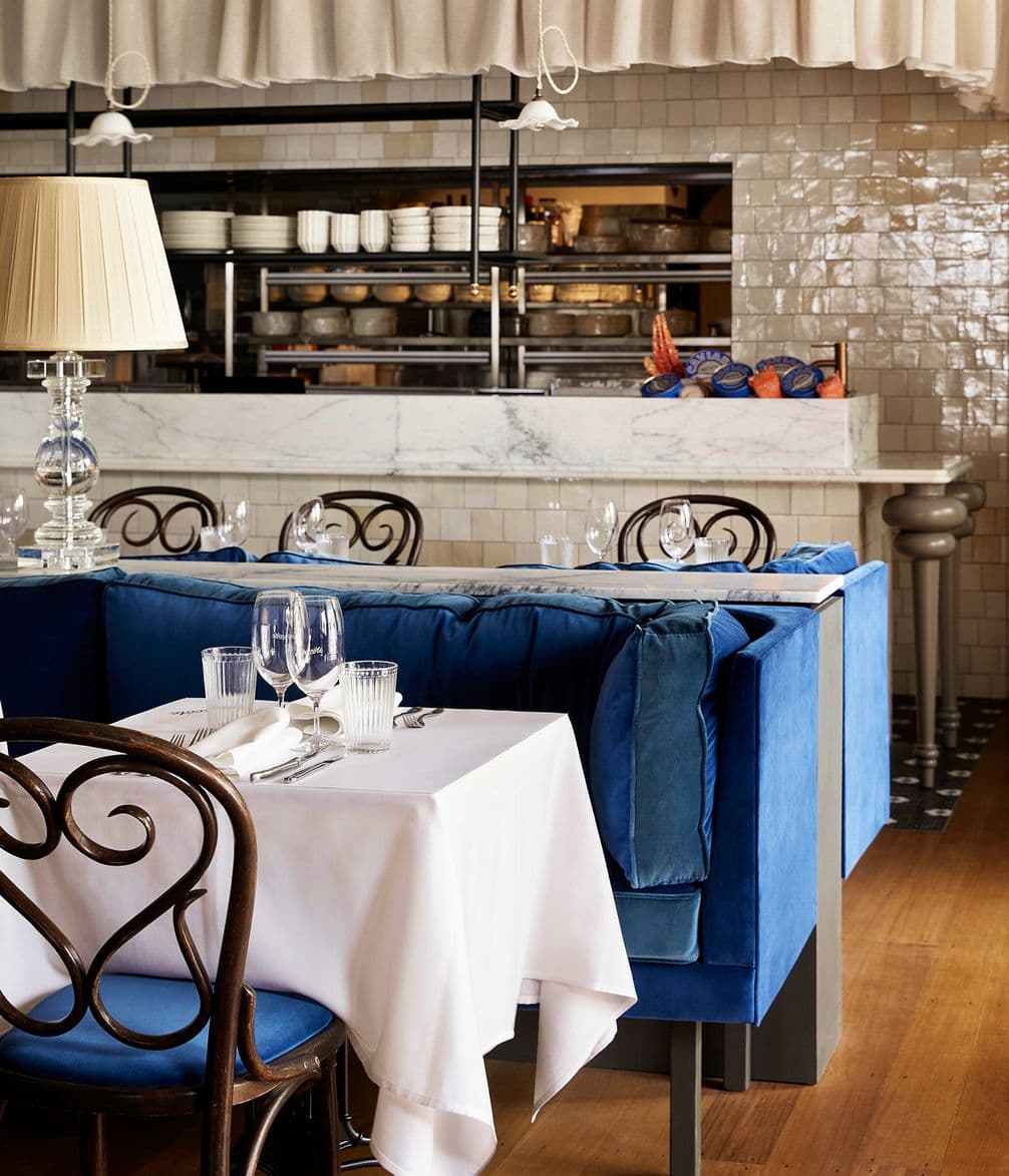 Elegant restaurant interior with blue velvet seating, white tablecloths, set tables, ornate chairs, and an open kitchen with a marble counter.
