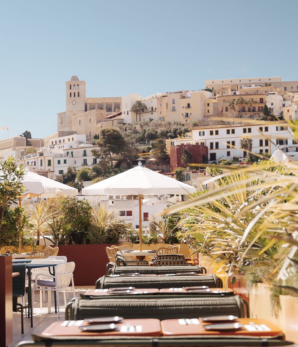 Outdoor cafe with tables and chairs, under white umbrellas, overlooks a hillside town with beige buildings and a prominent stone tower under a clear sky.