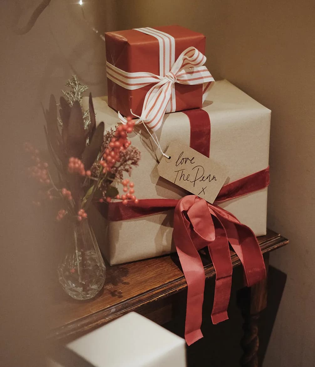 Two wrapped gifts with red ribbons sit on a table beside a small vase of flowers and berries. A note reads "Love The Pan."