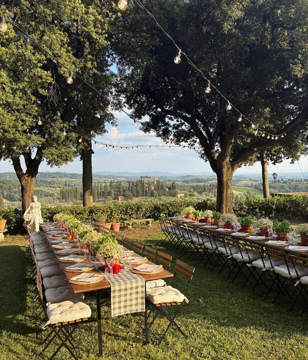 Outdoor dining tables are set for a meal under string lights, overlooking a scenic Italian landscape with vineyards and distant buildings.