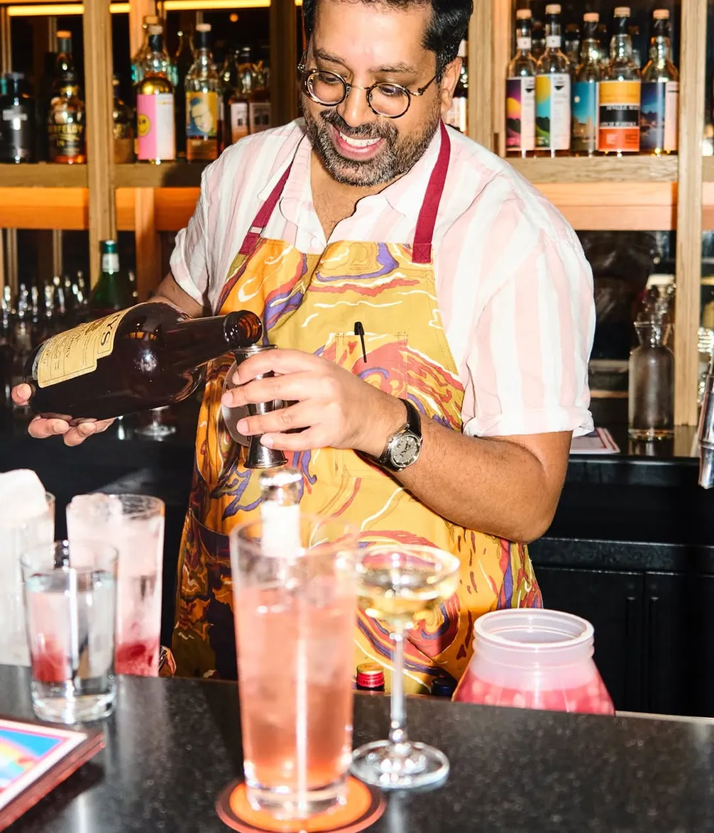 A bartender in a colorful apron is smiling while pouring a drink behind a bar, surrounded by various bottles and cocktail glasses.