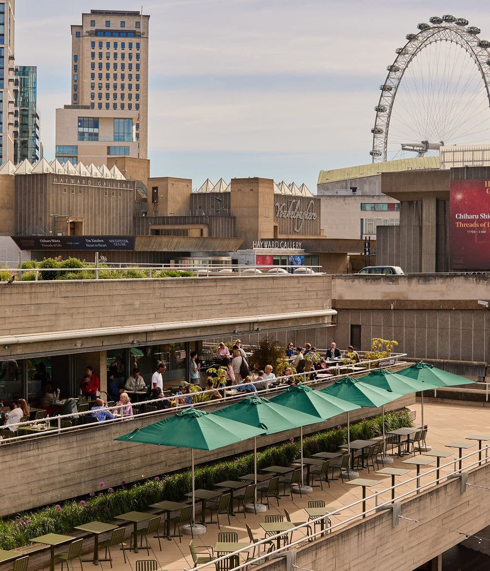 An outdoor café terrace with green umbrellas on a concrete building, city skyline, and a large Ferris wheel in the background.