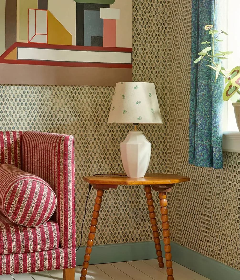 A cozy room corner with a striped red couch, wooden side table, geometric lamp, patterned wallpaper, abstract wall art, and blue curtain by a window.
