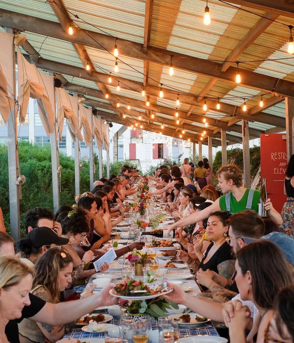 A long communal table under a canopy with people dining together. String lights hang overhead, and a server is handing a dish to a guest.